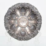 Fruteira em metal prateado, formato circular, com bojo raso e bordo recortado; centro decorado em relevo por repuxo, com composição radial de reservas lisas alternadas com conchas estilizadas, envolvidas por motivos de volutas, elementos fitomorfos e pequenas rosetas. Assentado sobre base circular baixa, levemente escalonada. Necessita refazer banho de prata. Medidas: