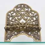 Suporte para bíblia em metal fundido vazado, painel frontal recortado com volutas e folhas de acanto, base com apoio inferior e haste posterior articulada para regulagem de inclinação, barra frontal para retenção do livro. Medidas: