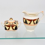 J & G Meakin, Staffordshire - Inglaterra(Fundada em 1851). Uma(01) leiteira e um(01) açucareiro em faiança inglesa da década de 1930, padrão "SOL"; esmaltação de tom marfim, dotado de rica decoração Art Déco, contendo composição geométrica radial, formada por reservas triangulares alternadas em esmalte de tonalidade preta, marrom e amarelo-creme, dispostas em sequência contínua, bojo circundado por frisos concêntricos em tom laranja e mostarda, este último se replica no contorno da borda da tampa. Marca da manufatura no verso. Medidas: Leiteira: Alt.: 14,5 x Comp.: 16 x Larg.: 11 cm; Açucareiro: Alt.: 9,5 x Comp.: 14,5 x Larg.:  9 cm.