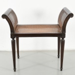 Banqueta construída em madeira, de formato retangular, com assento e espaldar duplo em trama de palhinha natural, laterais compostas por montantes curvos em voluta formando apoios elevados, unidos por travessa superior cilíndrica, estrutura assentada sobre quatro pernas torneadas e afiladas, com caneluras verticais e terminações simples, ligadas por longarinas retas. Elementos de ligação com blocos decorativos esculpidos em motivo geométrico. Estado de conservação conforme fotografias, apresenta sinais de uso. Medidas: Alt.: 62 x Comp.: 68 x Larg.: 37 cm.