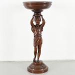 Floreira de coluna / porte-plante em madeira maciça entalhada, com base circular escalonada, decorada por friso em relevo com motivos fitomorfos. Fuste escultórico em forma de cariátide feminina, de vulto pleno, em posição frontal, com braços elevados sustentando elemento superior, trajando túnica de inspiração clássica com panejamento definido. Capitel configurado em taça circular côncava, com borda espessada e perfil moldurado. Medidas: Alt.: 91 x Diam.: 37 cm.