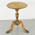 Mesa lateral / guéridon em madeira entalhada e dourada, com tampo circular de bordo moldurado, fuste central torneado com seção canelada e base trípode com pernas em voluta, terminadas em pés zoomórficos estilizados. Decoração com elementos de inspiração clássica, incluindo caneluras e volutas. Acabamento em douração com pátina e desgaste visível. Medidas: Alt.: 49 x Diam.: 36 cm.