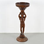 Floreira de coluna / porte-plante em madeira maciça entalhada, com base circular escalonada, decorada por friso em relevo com motivos fitomorfos. Fuste escultórico em forma de cariátide feminina, de vulto pleno, em posição frontal, com braços elevados sustentando elemento superior, trajando túnica de inspiração clássica com panejamento definido. Capitel configurado em taça circular côncava, com borda espessada e perfil moldurado. Medidas: Alt.: 98 x Diam.: 35,5 cm.