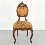 Cadeira em madeira entalhada, em estilo Luís XV (rocaille), com espaldar de formato ovalado vazado, encimado por ornato em volutas, concheados e motivos florais em relevo, ladeado por montantes curvos. Assento de contorno serpenteado, estofado, revestido em tecido com padrão floral estilizado em tons de bege e dourado, arrematado por galão perimetral na tonalidade dourada. Cintura recortada com decoração entalhada de elementos fitomorfos e concheados, apoiando-se sobre quatro pernas, sendo as dianteiras em curva cabriolet com joelho ornado e pés em voluta, e as traseiras recuadas de secção mais simples. Madeira em tonalidade castanho médio a escuro, com pátina e desgastes compatíveis com o uso. Medidas: Alt.: 103 x Comp.: 51,5 x Larg.: 42 cm.