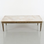 Mesa de centro em madeira entalhada e dourada, de estilo neoclássico, com tampo retangular em mármore. Saia decorada com frisos geométricos e cartela central com motivos vegetalistas em relevo. Sustentação por quatro pernas torneadas, com caneluras verticais e elementos ornamentais na transição para a cintura. Medidas: Alt.: 35 x Comp.: 118 x Larg.: 54,5 cm.