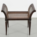 Banqueta construída em madeira, em estilo neoclássico de influência inglesa (Sheraton), com estrutura retangular e assento em palhinha natural trançada. Encostos laterais em forma de voluta contínua, com painéis em palhinha, configurando braços integrados. Estrutura sustentada por quatro pernas torneadas de secção circular, levemente afiladas, com caneluras verticais, unidas por travessas longitudinais e transversais. Elementos de ligação com entalhes geométricos simplificados nos cantos. Apresenta pequenas avarias, demanda restauro. Medidas: Alt.: 61 x Comp.: 69 x Larg.: 38 cm.