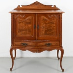 Móvel bar construído em madeira de tom claro, de influência do estilo Luís XV, com linhas curvas, pernas cabriolé e ornamentação com motivos fitomorfos em baixo-relevo; corpo com duas portas envidraçadas, interior espelhado e prateleiras em vidro, além de dois gavetões inferiores alinhados; ferragens em metal vazado; remate superior recortado com elemento central. Parte de trás apresenta vestígio xilófago. Medidas: