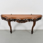 Console de parede construído em madeira entalhada, com tampo recortado em contorno ondulado e borda moldurada em ressaltos sucessivos. Saia frontal vazada, ricamente esculpida com volutas, concheados e motivos fitomórficos, organizados em composição simétrica, com elemento central em destaque. Laterais igualmente vazadas, integrando-se ao desenho da travessa. Apresenta duas pernas em cabriolet, de perfil arqueado, com joelhos marcados por entalhes e pés em voluta, decorada por filetes e elementos geométricos incisos. Estrutura com travamento inferior integrado à própria saia entalhada, dispensando travessas adicionais aparentes. Fixação posterior com argolas metálicas para afixação em parede. Medidas: Alt.: 55 x Comp.: 104 x Larg.: 50 cm.