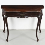 Mesa de encostar em madeira, com tampo recortado de contorno sinuoso, moldurado, sobre caixa com uma gaveta frontal. Saia ondulada com entalhe central em motivo floral e puxador metálico fundido de desenho vazado. Apoio sobre quatro pernas cabriolet, com joelhos entalhados em volutas e terminação em pés curvos. Originalmente com tampo duplo, perdido neste exemplar. Medidas: Alt.: 79 x Comp.: 107 x Larg.: 51 cm.