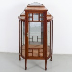 Excepcional vitrine em madeira nobre de tom claro, decorada com faixas marchetadas. Parte superior dotada de espelhos bisotados, com moldura de recorte ondulado fixados por garras em bronze, tampos em mármore com rajados. Corpo central retangular de seção reta, dotado de porta frontal, e laterais bombée, emoldurados com placas em cristal bisotado. Interior contendo três prateleiras de vidro e fundo espelhado, assente sobre quatro pernas retilíneas afinando para baixo. Med.: Alt.: 151 x Comp.: 76 x Larg.: 33 cm.