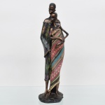 Escultura em resina moldada com pátina em tonalidade bronzeada, representando casal em composição vertical, com figuras estilizadas de proporções alongadas. A figura masculina posiciona-se posteriormente, envolvendo a feminina, que apresenta ventre proeminente, indicando gestação. Vestimentas com aplicação de tecido em drapeado contínuo, com padrão listrado e elementos florais.  Medidas: Alt.: 41 x Comp.: 11,5 x Larg.: 11 cm..