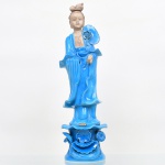 Escultura de Guanyin (Avalokitevara), executada em cerâmica esmaltada, forma sinizada do bodhisattva Avalokitevara, representada em posição ereta sobre base modelada com elementos de lótus estilizado, vestindo túnica de amplas mangas com pregueado vertical, com cabeleira disposta em coque alto (ushnisha estilizada) e adorno superior. Atributo floral junto ao ombro. Superfície com vidrado monocromático azul, com áreas em reserva. Mão direita removível. Estado conforme fotografias. Apresenta quebradura na base. Medidas: 