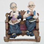 Escultura em resina moldada e policromada, representando casal de idosos sentado em banco. Grupo escultórico com duas figuras em composição única, apoiadas sobre banco com encosto recortado. Figura feminina trajando vestido com avental, segurando cesta com novelo de lã e instrumento de tricô; figura masculina trajando camisa e colete, portando cachimbo e livro. Fisionomias caricaturais, com aplicação de óculos em relevo. Policromia aplicada sobre base clara, com variações tonais nas vestes e detalhes. Medidas