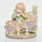 Escultura em resina policromada representando criança sentada em leito, em composição narrativa doméstica, com vestes em tons pastéis e elementos têxteis simulados (almofada e colcha), base oval integrada ao conjunto. Medidas: