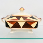 J & G Meakin, Staffordshire - Inglaterra(Fundada em 1851). Sopeira em faiança inglesa da década de 1930, padrão "SOL". Formato quadrangular, de quinas levemente chanfradas, bordo recortado em ondulações, tampa ligeiramente abaulada, puxador com feitio de pião, alças laterais curvadas, vazadas e modeladas em forma de concha estilizada, assente sobre quatro pés levemente revirados; esmaltação de tom marfim, dotado de rica decoração Art Déco, contendo composição geométrica radial, formada por reservas triangulares alternadas em esmalte de tonalidade preta, marrom e amarelo-creme, dispostas em sequência contínua. Centro e borda inferior circundados por frisos concêntricos em tom laranja e mostarda, este último se replica no contorno da borda da tampa. Marca da manufatura no verso. Medidas: Alt.: 18 x Comp.: 29 x Larg.: 22 cm. Estado de conservação conforme fotografias. Apresenta desgastes pontuais de uso, vide foto.