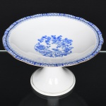 ROSSLAU - ALEMANHA. Exuberante fruteira de pedestal em porcelana alemã, formato circular de interior ligeiramente côncavo, contorno recortado com aba larga levemente ondulada, apoiado sobre base alta de formato cônico. Esmaltação branca, decoração apresenta composição em azul padrão china-blau, característica da tradição decorativa centro-europeia inspirada nas porcelanas orientais; aba ornada por friso contínuo em azul com motivos florais estilizados e elementos geométricos, delimitado por filete dourado acompanhando o contorno ondulado da borda, campo central apresenta vinheta decorativa em azul, composta por arranjo floral acompanhado de pequeno elemento ornamental representando ânfora sobre base. Marca na base em azul: CHINA-BLAU / ROSSLAU, indicando manufatura da fábrica Rosslau, Alemanha. Medidas: Alt.: 15 x Diam.: 25 cm.