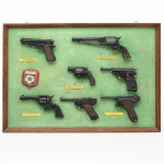 MILITARIA. Quadro expositivo com conjunto de miniaturas de armas de fogo, fixadas sobre fundo revestido em tecido de cor verde, acondicionado em moldura retangular em madeira. Composição organizada com exemplares dispostos em alinhamento horizontal e vertical, acompanhados por plaquetas identificadoras metálicas, com inscrições como Colt .45 Auto M1911A1, Remington N.M. Army .44, Smith & Wesson .38, Colt Frontier .44-40 e Luger P.08. Inclui elemento heráldico aplicado, representando insígnia com a inscrição Brasil.Miniaturas em metal com empunhaduras em material sintético ou madeira, de caráter decorativo, fixadas por suportes metálicos.