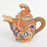 Excepcional bule em porcelana japonesa, decoração policroma, aplicação moriage em relevo, generosos arremates em pintura à ouro; bojo oval ornamentado com composição figurativa, formada por deidade central sentada, acompanhada por quatro Imortais(sennin/rakan), representando anciãos em trajes tradicionais, tema recorrente em produções de exportação em estilo Satsuma, fundo preenchido por padrão de ondas, com áreas delimitadas por contornos dourados. Pega da tampa, bico vertedor e alça lateral modelados em forma de dragão, dando a impressão do dragão estar atravessando o bojo de um lado para o outro. A iconografia e a técnica de moriage situam a peça dentro da produção japonesa voltada ao mercado externo entre o final do período Taisho e o início do período Showa. Fundo apresenta marca de manufatura com caracteres japoneses em vermelho-ferro. Medida: Alt.: 16 x Comp.: 18 x Larg.: 10 cm. Puxador da tampa com a cabeça colada.