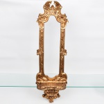 Exuberante espelho de parede com console integrado, construído em madeira entalhada e dourada, estilo Luís XV (rococó), de composição vertical com moldura recortada em contorno movimentado, ornamentada por volutas, concheados e elementos fitomórficos em relevo, de caráter rocaille; base com pequena mísula projetada, de superfície reticulada, destinada ao apoio de objetos ou imagem devocional, sustentada por elemento inferior em forma de concha estilizada; verso com suporte metálico para fixação e vestígios de etiqueta comercial. Necessita colocar vidro, perdido neste exemplar. Medidas: