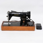 Máquina de costura elétrica, marca Singer, modelo 15C, fabricada no Brasil. Corpo em ferro fundido com acabamento em pintura esmaltada de tonalidade preta e decoração aplicada em dourado. Base em caixa de madeira revestida, com compartimento lateral para acessórios.Equipada com volante lateral, alavanca de controle de ponto com graduação numérica, placa de agulha em metal, sistema de lançadeira oscilante e acionamento por pedal elétrico externo. Iluminação integrada com lâmpada frontal protegida por capa metálica. Motor elétrico acoplado, identificado como Singer Simanco, com especificação de alimentação elétrica: 100110V, 2575 ciclos, potência aproximada de 0,55 A. Identificações de fábrica e numeração presentes no corpo e no motor. Estado de conservação conforme fotografias.