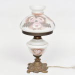 Abajur de mesa em metal fundido e vidro opalinado; base em bronze com fundição em relevo composta por volutas e motivos vegetalistas, haste central com colar metálico, reservatório com decoração floral aplicada, cúpula em forma de campânula com decoração floral pintada e friso dourado, dotada de abertura superior destinada à ventilação térmica do conjunto elétrico; estado de conservação com desgaste do acabamento metálico com oxidação, sujidade aderida, pintura do vidro com perdas pontuais. Necessita refazer parte elétrica.  Medidas: