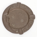 Cinzeiro em metal fundido com representação de face lunar e estrelas, provável liga de cobre (bronze), moldagem em relevo, sem marcas de fabricante aparentes, de linguagem Art Nouveau tardia, primeira metade do século XX; corpo circular com três apoios equidistantes para repouso de cigarro, aba externa ornamentada com motivos vegetalistas estilizados, campo central texturizado com cinco estrelas vazadas, lateral com representação de lua minguante com face humana em perfil integrada à borda, verso com rebaixo circular e pontos de apoio; estado de conservação: desgaste da superfície, pátina irregular com áreas de oxidação, resíduos aderidos e pontos de abrasão. Medidas: