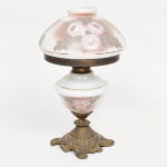 Abajur de mesa em metal fundido e vidro opalinado; base em bronze com fundição em relevo composta por volutas e motivos vegetalistas, haste central com colar metálico, reservatório com decoração floral aplicada, cúpula em forma de campânula com decoração floral pintada e friso dourado, dotada de abertura superior destinada à ventilação térmica do conjunto elétrico; estado de conservação com desgaste do acabamento metálico com oxidação, sujidade aderida, pintura do vidro com perdas pontuais, fissura no colo da cúpula. Necessita refazer parte elétrica.  Medidas: