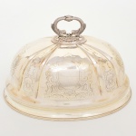 Cloche (cobertura de serviço) para carne/assados confeccionado em metal prateado, formato oblongo de perfil abaulado, moldado com frisos verticais e campos gravados com cartelas rocaille e volutas; pega superior fundida em forma de golfinhos afrontados, com texturização de escamas e base circular perlada em relevo, obtidos através da técnica do cinzelado; bordo inferior com filetes concêntricos. Ausência de marca de manufatura. Medidas: Alt.:23,5 x Comp.: 36 x Larg.: 26 cm. Apresenta pequenos desgastes isolados no banho de prata(sangria), necessita polimento.