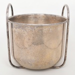 WOLFF. Champanheira em metal prateado, de corpo circular com paredes ligeiramente inclinadas e borda lisa. Estrutura com armação externa em hastes metálicas, formando suportes laterais elevados com alças em arco integradas. Marca da manufatura ao fundo. Necessita polimento /ou refazer banho de prata. Medidas: