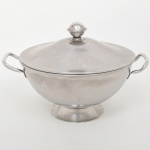 Sopeira com tampa confeccionada em aço inoxidável, corpo hemisférico com bordo liso, assente sobre pé circular alargado; duas alças laterais em arco, aplicadas; tampa abobadada com pega esférica central. Marca Luralite Inox gravada na base. Medidas: 21,5 x Comp.: 33 x Larg.: 26 cm.