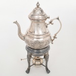 Chocolateira (chocolatière) com trempe confeccionada em metal espessurado à prata; corpo bojudo com cintura marcada e base anelada, decorado em relevo com motivos vegetalistas estilizados; bico vertedor recurvo com extremidade revirada para fora, asa lateral em voluta, tampa campaniforme, dotada de abertura basculante, encimada por pega com formato de pináculo. Conjunto acompanhado de trempe tripé com pés recortados e reservatório inferior para aquecimento, dotado de queimador. Apresenta desgastes parciais no banho de prata, e necessita polimento. Medidas: Alt.: 39 x Comp.: 25 x Larg.: 13 cm.