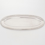 Bandeja oval confeccionada em metal prateado, centro ligeiramente rebaixado, borda relevada com frisos e folhagens. Medidas: