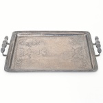 Bandeja confeccionada em metal prateado, formato retangular de quinas arredondadas, centro ligeiramente rebaixado, lavrado com composição de motivo vegetalista, borda de contorno relevado com fileira de frisos e fitas cruzadas, alças laterais fusiformes apoiadas sobre hastes curvas. Necessita polimento e/ou refazer banho de prata. Medidas: