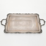 REGIA PRATA 90. Elegante bandeja confeccionada em metal espessurado à prata, formato retangular de quinas arredondadas, centro ligeiramente rebaixado, ricamente lavrado com composição de motivo vegetalista, borda relevada com volutas e conchas, ornato replicado nas alças laterais. Marca da manufatura no verso. Necessita polimento e/ou refazer banho de prata. Medidas: