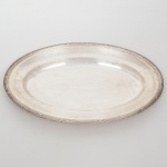 FRACALANZA. Elegante bandeja confeccionada em metal espessurado à prata, formato oval, centro ligeiramente rebaixado, borda relevada com fileira de frisos intercaladas por fitas cruzadas, ornato obtido através da técnica do cinzelado. Marca da manufatura no verso. Medidas: