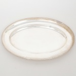 WOLFF. Elegante bandeja confeccionada em metal espessurado à prata, formato oval, centro ligeiramente rebaixado, borda relevada com fileira de frisos intercaladas por fitas cruzadas, ornato obtido através da técnica do cinzelado. Marca da manufatura no verso. Medidas:
