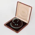 BIJUTERIA. Parure composta por colar, pulseira e par de brincos em metal prateado, com armações articuladas, cravação de pedras de tonalidade verde esmeralda, lapidadas em diferentes cortes (retangular, pera e circular) intercaladas por pedraria incolor; colar de conformação semicircular com elementos pendentes; pulseira em estrutura flexível de duas fileiras; brincos circulares com pedra central facetada envolta por halo de pedras menores. Acondicionada em estojo original. Apresenta desgastes do prateado, uma perda da pedraria incolor do colar e pequeno sinal de oxidação. Medidas: Colar(fechado): 18 cm. Pulseira: Comp.: 6,5 cm. Brincos: Diam.: 1,8 cm.