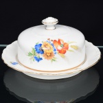 Springer & Co, EPIAG - Checoslováquia(Fundada entre 1918 e 1920 a partir de fábricas nos arredores de Carlsbad). Excepcional manteigueira em porcelana tcheca da década de 1920. Esmaltagem vítrea na cor branca, decorada com bela composição botânica em estampagem policromada, predominando as tonalidades amarelo, ocre, azul, coral, verde e marsala, dispostas em agrupamento lateral e pequenos ramos distribuídos pela tampa e aba do presentoir; orla do prato delimitada por faixa contendo frisos e enrolamentos relevados na própria pasta, borda recortada com contorno ornamentado com aplicação de filete em douradura; tampa abobadada encimada por puxador com feitio de pião. Marca da manufatura no verso. Medida: Alt.: 9,5 x Diam.: 18,5 cm. (A Springer & Co de Elbogen na Boêmia foi formada na década de 1880 e encerrou suas atividades em 1945. Ela foi uma das fábricas fundadoras originais da OEPIAG (mais tarde EPIAG), que era uma associação de olarias formadas pelo governo de Viena em 1918 para promover a venda de cerâmica e porcelana da Boêmia. Foi inicialmente denominado OEPIAG, um acrônimo para Österreichische Porzellan Industrie AG ou Austrian Porcelain Industry, mas foi alterado para EPIAG - Erste Böhemische Porzellan Industrie AG que significa Primeira Indústria de Porcelana da Boêmia - em 1920 após a independência da Checoslováquia).
