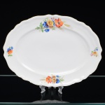 Springer & Co, EPIAG - Checoslováquia(Fundada entre 1918 e 1920 a partir de fábricas nos arredores de Carlsbad). Excepcional travessa oval em porcelana tcheca da década de 1920. Esmaltagem vítrea na cor branca, centro ligeiramente rebaixado, decorada com bela composição botânica em estampagem policromada, predominando as tonalidades amarelo, ocre, azul, coral, verde e marsala, distribuídas pela aba; orla delimitada por faixa contendo frisos e enrolamentos vegetalistas relevados na própria pasta; borda recortada, e ondulada, com contorno ornamentado com aplicação de filete em douradura. Marca da manufatura no verso. Medidas: Comp.: 34,5 x Larg.: 26,5 cm. (A Springer & Co de Elbogen na Boêmia foi formada na década de 1880 e encerrou suas atividades em 1945. Ela foi uma das fábricas fundadoras originais da OEPIAG (mais tarde EPIAG), que era uma associação de olarias formadas pelo governo de Viena em 1918 para promover a venda de cerâmica e porcelana da Boêmia. Foi inicialmente denominado OEPIAG, um acrônimo para Österreichische Porzellan Industrie AG ou Austrian Porcelain Industry, mas foi alterado para EPIAG - Erste Böhemische Porzellan Industrie AG que significa Primeira Indústria de Porcelana da Boêmia - em 1920 após a independência da Checoslováquia).