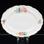 Springer & Co, EPIAG - Checoslováquia(Fundada entre 1918 e 1920 a partir de fábricas nos arredores de Carlsbad). Excepcional travessa oval em porcelana tcheca da década de 1920. Esmaltagem vítrea na cor branca, centro ligeiramente rebaixado, decorada com bela composição botânica em estampagem policromada, predominando as tonalidades amarelo, ocre, azul, coral, verde e marsala, distribuídas pela aba; orla delimitada por faixa contendo frisos e enrolamentos vegetalistas relevados na própria pasta; borda recortada, e ondulada, com contorno ornamentado com aplicação de filete em douradura. Marca da manufatura no verso. Medidas: Comp.: 31 x Larg.: 23,5 cm. (A Springer & Co de Elbogen na Boêmia foi formada na década de 1880 e encerrou suas atividades em 1945. Ela foi uma das fábricas fundadoras originais da OEPIAG (mais tarde EPIAG), que era uma associação de olarias formadas pelo governo de Viena em 1918 para promover a venda de cerâmica e porcelana da Boêmia. Foi inicialmente denominado OEPIAG, um acrônimo para Österreichische Porzellan Industrie AG ou Austrian Porcelain Industry, mas foi alterado para EPIAG - Erste Böhemische Porzellan Industrie AG que significa Primeira Indústria de Porcelana da Boêmia - em 1920 após a independência da Checoslováquia).