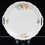 Springer & Co, EPIAG - Checoslováquia(Fundada entre 1918 e 1920 a partir de fábricas nos arredores de Carlsbad). Excepcional bandeja em porcelana tcheca da década de 1920. Esmaltagem vítrea na cor branca, decorada com bela composição botânica em estampagem policromada, predominando as tonalidades amarelo, ocre, azul, coral, verde e marsala, dispostas em agrupamento lateral e pequenos ramos distribuídos pela aba; orla delimitada por faixa contendo frisos e enrolamentos relevados na própria pasta; alças laterais integradas, borda recortada com contorno ornamentado com aplicação de filete em douradura. Marca da manufatura no verso. Medida: Diam.: 29 cm. (A Springer & Co de Elbogen na Boêmia foi formada na década de 1880 e encerrou suas atividades em 1945. Ela foi uma das fábricas fundadoras originais da OEPIAG (mais tarde EPIAG), que era uma associação de olarias formadas pelo governo de Viena em 1918 para promover a venda de cerâmica e porcelana da Boêmia. Foi inicialmente denominado OEPIAG, um acrônimo para Österreichische Porzellan Industrie AG ou Austrian Porcelain Industry, mas foi alterado para EPIAG - Erste Böhemische Porzellan Industrie AG que significa Primeira Indústria de Porcelana da Boêmia - em 1920 após a independência da Checoslováquia).