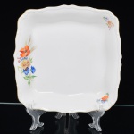 Springer & Co, EPIAG - Checoslováquia(Fundada entre 1918 e 1920 a partir de fábricas nos arredores de Carlsbad). Excepcional saladeira em porcelana tcheca da década de 1920. Esmaltagem vítrea na cor branca, formato quadrangular de quinas arredondadas, centro rebaixado, decorada com bela composição botânica em estampagem policromada, predominando as tonalidades amarelo, ocre, azul, coral, verde e marsala, dispostas em agrupamento lateral e pequenos ramos distribuídos pela aba; orla delimitada por faixa contendo frisos e enrolamentos relevados na própria pasta; alças laterais integradas, borda recortada com contorno ornamentado com aplicação de filete em douradura. Marca da manufatura no verso. Medida: Alt.: 5 x Comp.: 24,5 x Larg.: 24,5 cm. (A Springer & Co de Elbogen na Boêmia foi formada na década de 1880 e encerrou suas atividades em 1945. Ela foi uma das fábricas fundadoras originais da OEPIAG (mais tarde EPIAG), que era uma associação de olarias formadas pelo governo de Viena em 1918 para promover a venda de cerâmica e porcelana da Boêmia. Foi inicialmente denominado OEPIAG, um acrônimo para Österreichische Porzellan Industrie AG ou Austrian Porcelain Industry, mas foi alterado para EPIAG - Erste Böhemische Porzellan Industrie AG que significa Primeira Indústria de Porcelana da Boêmia - em 1920 após a independência da Checoslováquia).