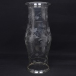 Donzela em vidro incolor, corpo alongado com leve constrição central, boca alargada com borda revirada e base circular com reservatório inferior para vela. Execução em vidro soprado em molde, com acabamento de bordo ao fogo. Decoração gravada composta por motivos vegetalistas com ramos, folhas e flores estilizadas, dispostos em frisos contínuos. Medidas: Alt.: 55 x Diam.: 73 cm.