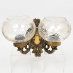 Arandela de parede confeccionada em bronze e metal amarelo, contendo duas luzes, braços em enrolamento vegetalista com extremo revirado para cima, cúpulas de formato globular achatado em meio cristal incolor, talhadas com motivos geométricos radiais estilizados, organizados com friso contínuo ao redor do bojo. Medidas: Lustre: Alt.: 22 x Comp.: 22 x Larg.: 15,5 cm; Cúpula: Alt.: 11 x Diam.: 14 cm.