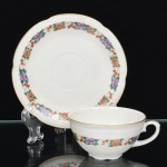 CHODZIEZ PORCELAIN, Chodziez - Polônia(Fundada em 1852 ou 1854, por Hermann Müller e Ludwik Schnorr). Xícara para café em porcelana polonesa da década de 1940; esmaltagem vítrea na cor branca, bojo hemisférico assentado sobre base circular recortada, decorada com faixas contendo padrão floral, aplicado em estampagem policromada, borda delimitada por barrado contínuo relevado na própria pasta, alça lateral em voluta com descanso de polegar relevado em folhagem, contornos realçados com filete em douradura, padrão decorativo replicado no pires. Marca da manufatura no verso. Medidas: Xícara: Alt.: 4,5 x Comp.: 9,5 x Larg.: 8 cm.