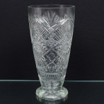 Vaso executado em meio cristal, diáfano e incolor, fomato obcônico assentado sobre base poligonal, ricamente talhado com sulcos, palmetas radiais, leques, estrelas e losangos cruzados, zona inferior apresenta faixa contínua de caneluras verticais (fluting), base facetada com treze lados, fundo preenchido por estrela de dezesseis pontas. Medidas: Alt.: 32 x Diam.: 16 cm.