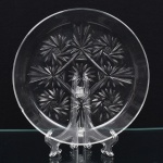 Prato em cristal europeu, diáfano e incolor, formato circular de borda elevada, decoração moldada em baixo relevo, organizada em composição radial geométrica, com predomínio de rosetas estelares (starburst) distribuídas em disposição losangular., incluindo estrela central multifacetada, circundada por estrelas secundárias de múltiplas pontas, além de elementos lanceolados irradiantes. Apresenta um bicado na borda, vide foto. Medidas: Alt.: 2,5 x Diam.: 22 cm.