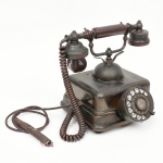 Antigo telefone de disco confeccionado em metal e outros materiais, receptor auricular em baquelite marrom, ligado por cabo espiralado. Haste superior com empunhadura central em material isolante (baquelite ou ebonite), suspensa por suportes metálicos. Sistema de discagem por mostrador rotativo frontal (rotary dial), com anel numerado (09) e indicações alfabéticas (ABC, DEF, GHI, etc.), padrão introduzido em aparelhos automáticos a partir da década de 1920. No estado, não foi testado, sem garantias. Fiação partida. Med.: Alt. 24 x Comp. 18 x Larg. 22,5 cm.