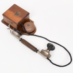 Siemens & Halske, fundada em 1847 em Berlim, pioneira no desenvolvimento de sistemas telegráficos e telefônicos, responsável pela difusão dos primeiros aparelhos telefônicos comerciais na Europa e posteriormente em diversos mercados internacionais. Telefone de parede, sistema magneto, com caixa em madeira, de formato retangular com tampa superior removível. O conjunto apresenta campainha externa metálica (timbre polarizado) acoplada à base, acionada por manivela lateral (gerador magneto), destinada à chamada manual em redes não automatizadas. Receptor tipo bastão (handset primitivo separado), com empunhadura em madeira torneada e cápsulas metálicas nas extremidades, conectado por fio revestido. Transmissor fixo em formato de corneta (bocal), típico dos primeiros sistemas telefônicos. Estrutura metálica com suporte de gancho para repouso do receptor. Placa frontal com emblema da Siemens, indicando produção original da companhia. Fabricação alemã, final do século XIX a início do século XX, circa 18951910. Não foi testado, sem garantias de funcionamento e estado do maquinário interno. Medidas: Caixa: Alt.: 21,5 x Comp.: 12,5 x Larg.: 8 cm. Receptor: Comp.: 29 cm.