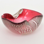 Centro de mesa em vidro artístico, com corpo em formato livre e borda ondulada irregular, apresentando cavidades moldadas por pinçamento manual; execução em vidro soprado, com aplicação de pó de prata (aventalina) sobre fundo rubi translúcido, criando efeito moteado. Apresenta acentuadas quebraduras, demanda grande restauro. Medidas: Alt.: 6 x Comp.: 14 x Larg.: 11 cm.